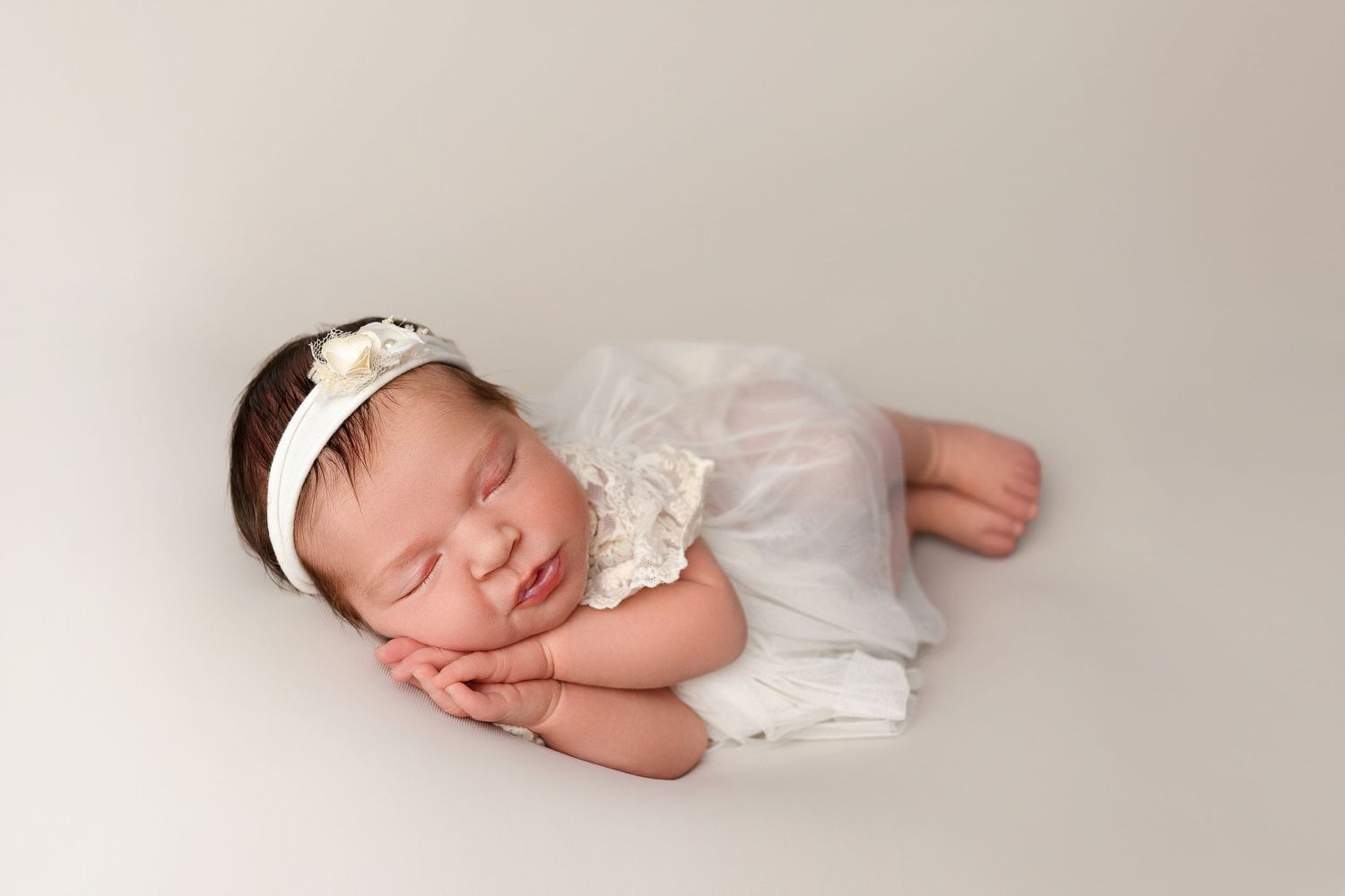 Sleeping newborn in white lace dress and headband rests peacefully on light backdrop during professional photoshoot.