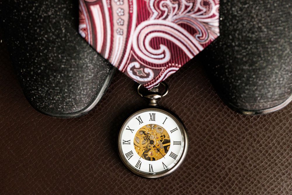 Vintage pocket watch with roman numerals displayed beside gray shoes and red tie.