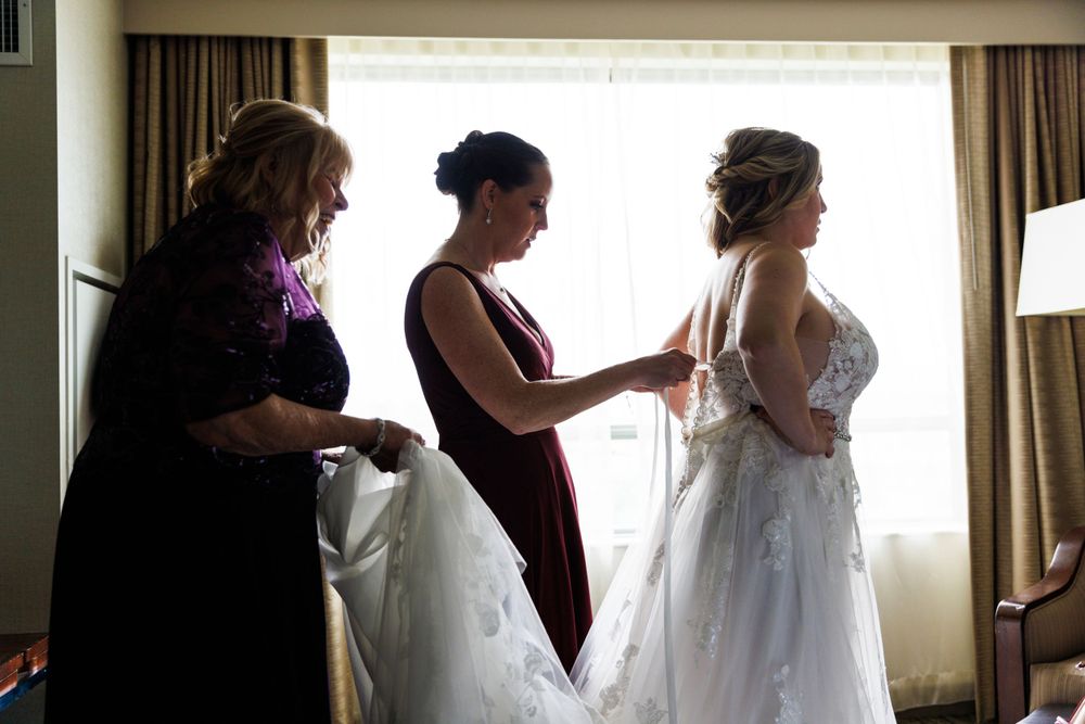 Bride being helped into wedding dress by two women in hotel room.