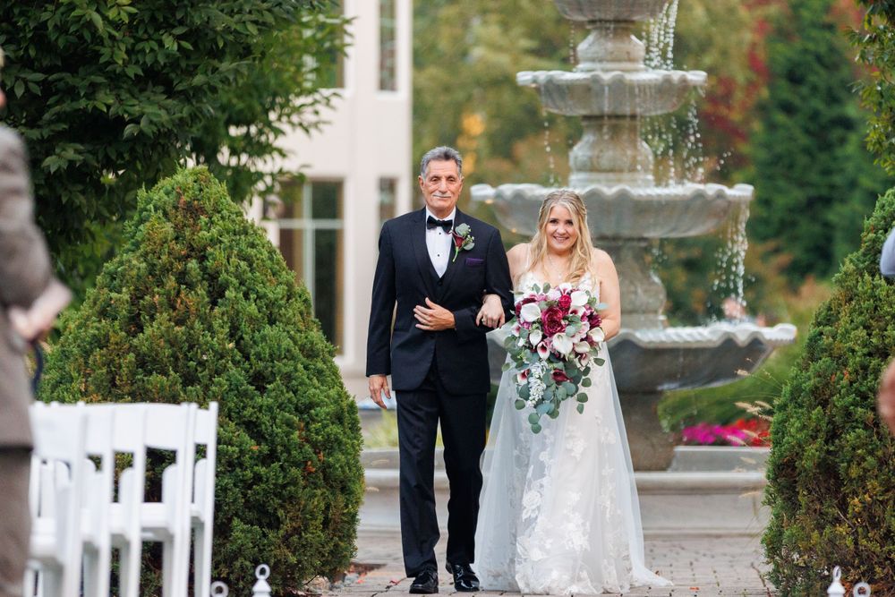 Father walks bride down outdoor aisle past fountain at elegant garden wedding.