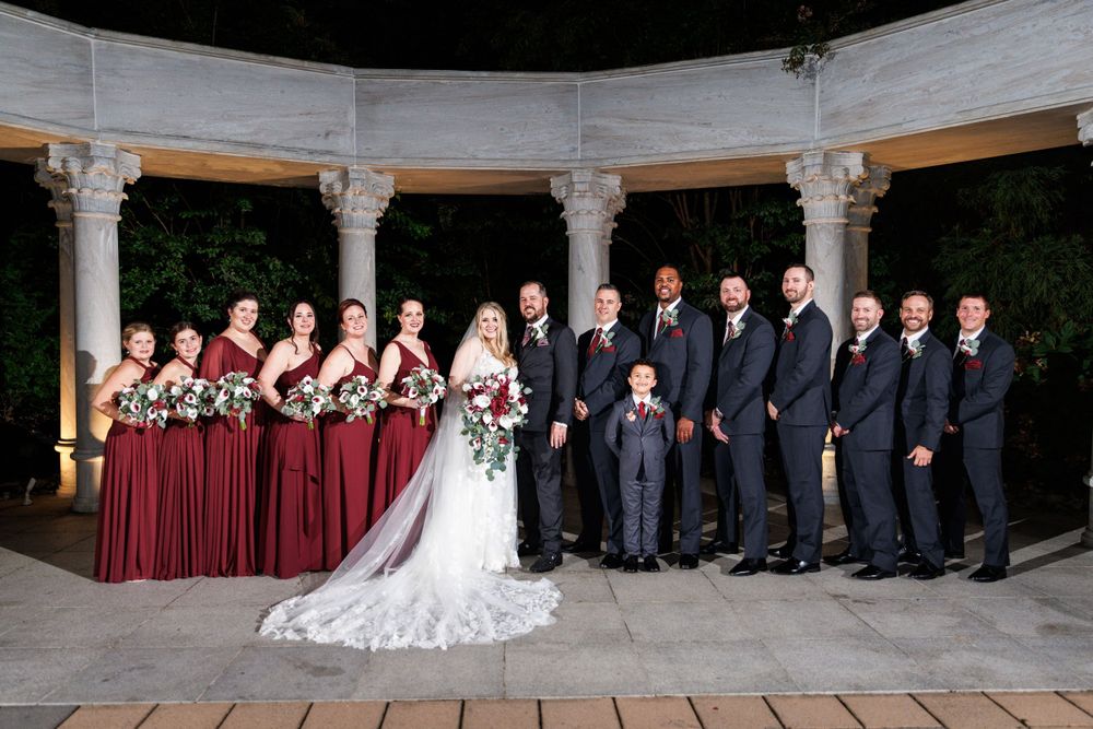 Wedding party posing under a stone rotunda, bridesmaids in burgundy gowns and groomsmen in navy suits.