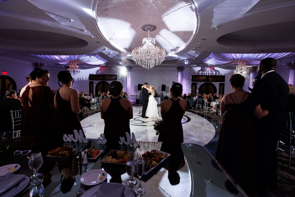 Elegant wedding reception with guests in black attire surrounding a couple sharing their first dance under a chandelier.