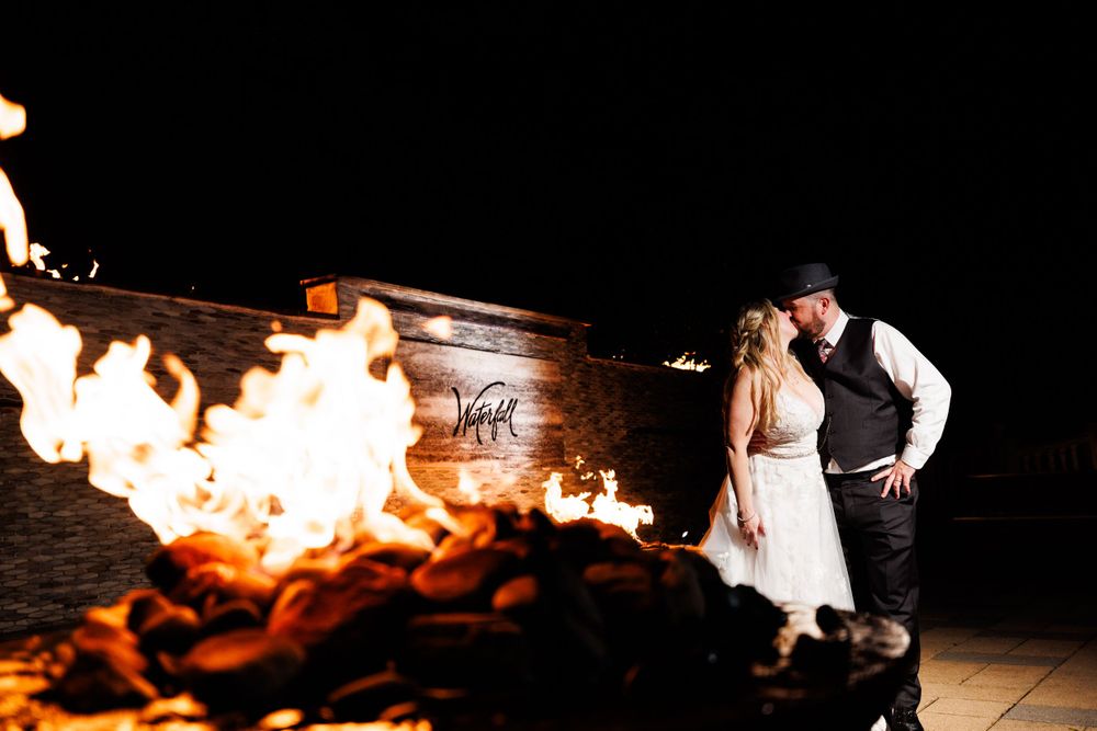 Newlyweds sharing a romantic kiss beside a roaring fire pit during their outdoor evening wedding celebration.