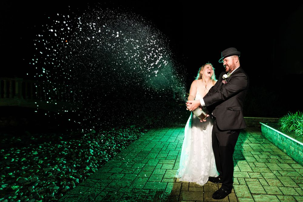 Bride and groom popping a champagne bottle outdoors at night with confetti bursting into the air.