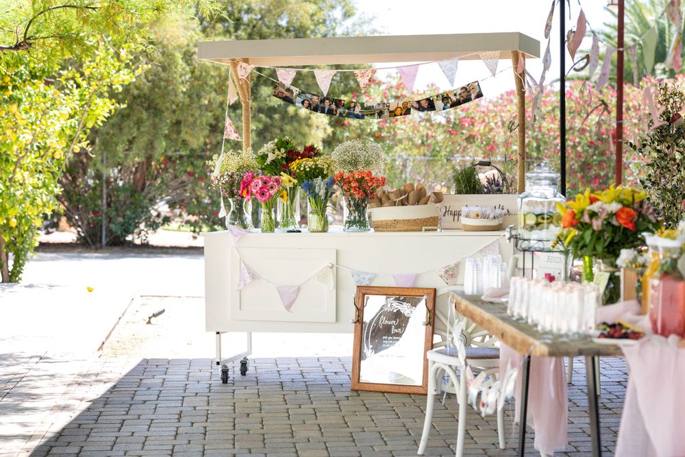 Rustic outdoor garden party setup with floral arrangements, string lights, and white furniture on a stone patio.