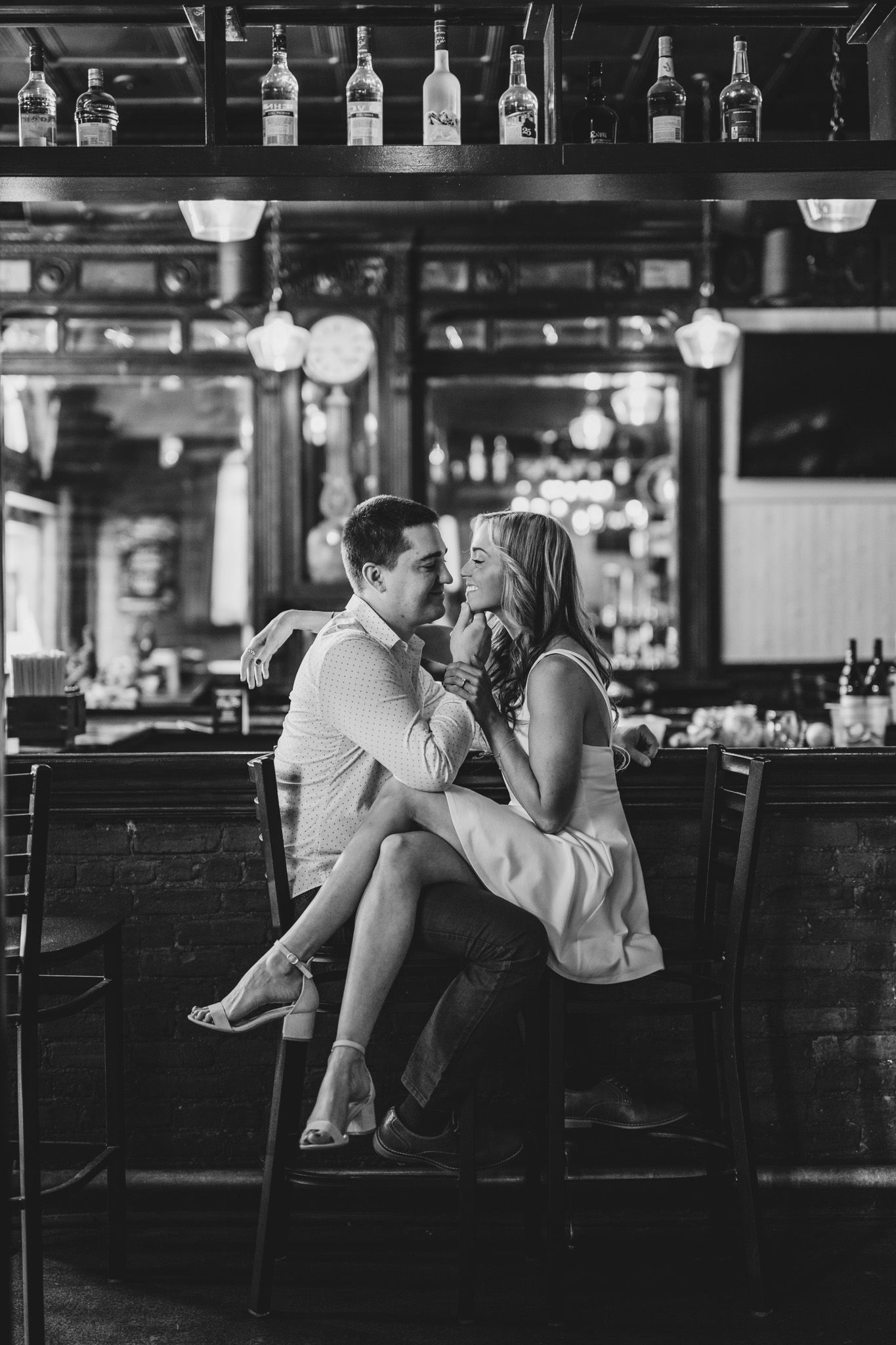 A romantic black and white photo of a couple sharing an intimate moment at a bar counter with bottles displayed above.