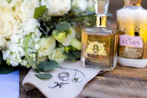 Wedding details with perfume bottle, rings, and white floral arrangement on wooden surface.