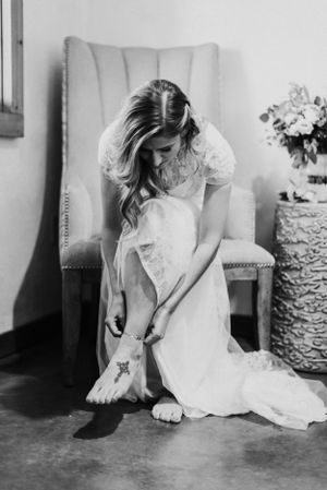 A black and white photo of a bride putting on her wedding shoes while seated in a tufted chair.