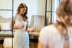 A bride in a fitted lace wedding dress looks at herself while getting ready in an elegant room.