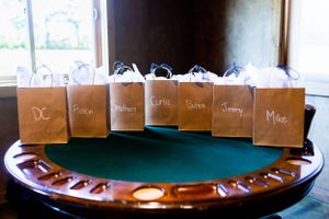 Brown paper gift bags labeled for the wedding party arranged on a green table.