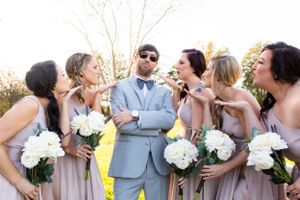 Wedding party in blush dresses and grey suit posing playfully with white floral bouquets.