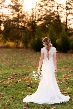 A backlit bride in a lace wedding dress stands in a field at sunset holding a bouquet.
