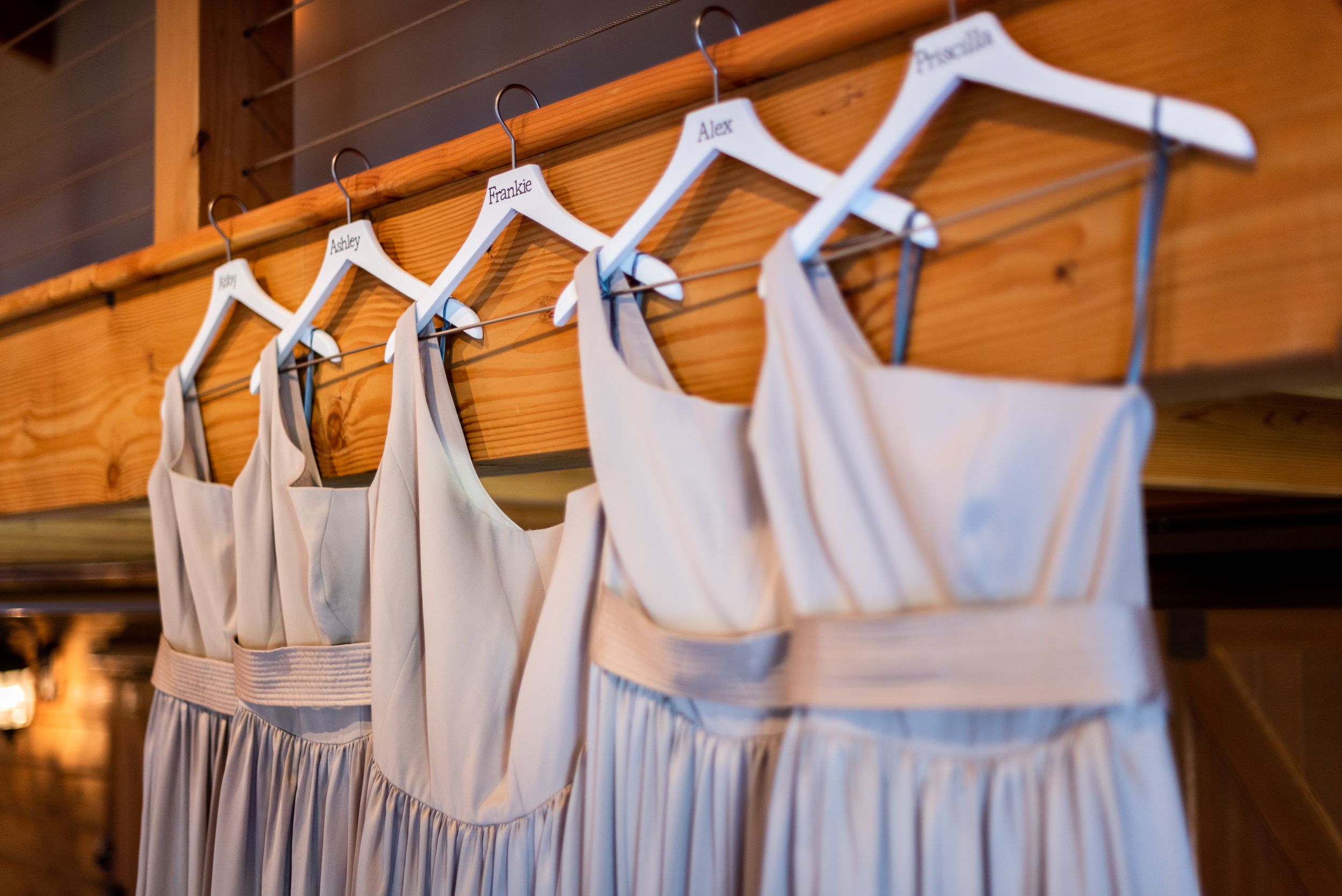 Row of light gray bridesmaid dresses hanging on white hangers in a rustic venue.