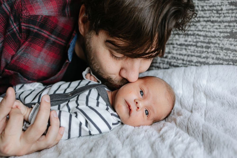 Baby wearing a striped onesie lies on white bedding while being lovingly held in a tender moment.