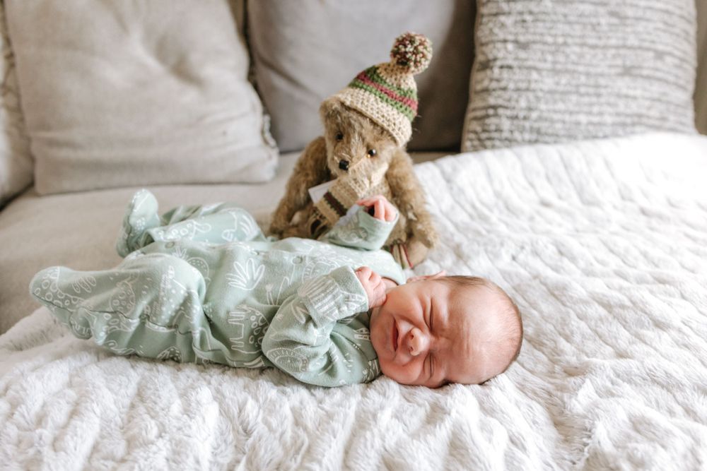 Golden dog wearing knit hat watches over baby in mint green swaddle sleeping on white bedding.