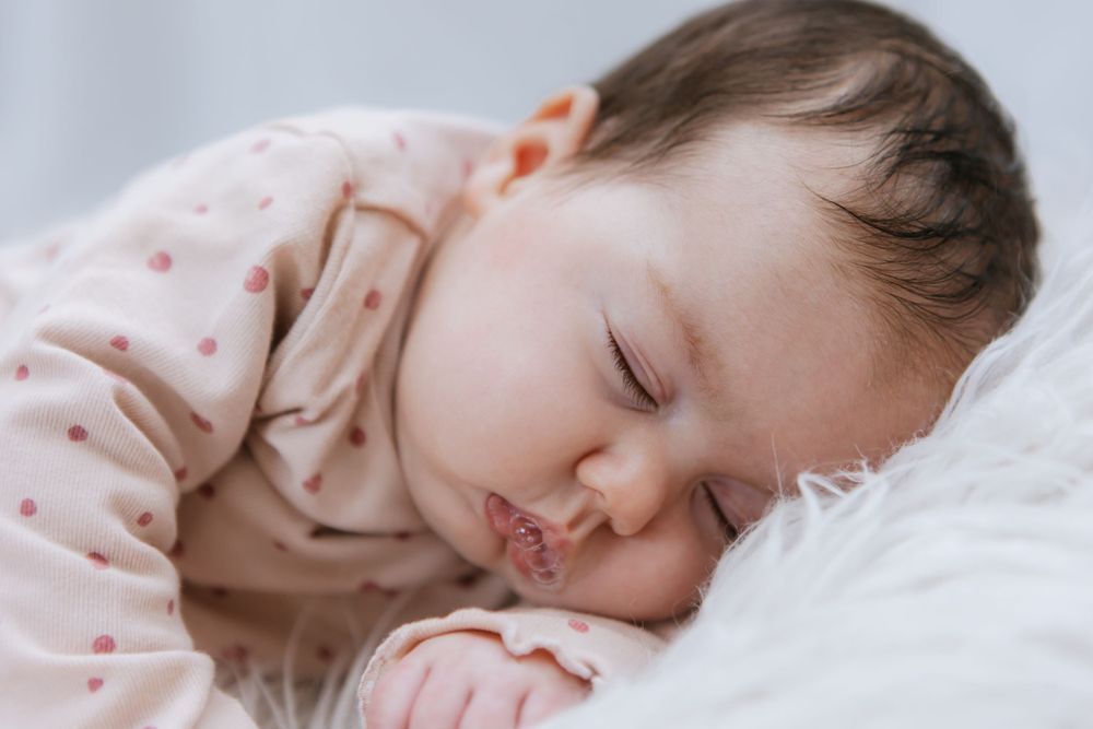 A sleeping baby wearing a pink polka dot outfit rests peacefully on white bedding.