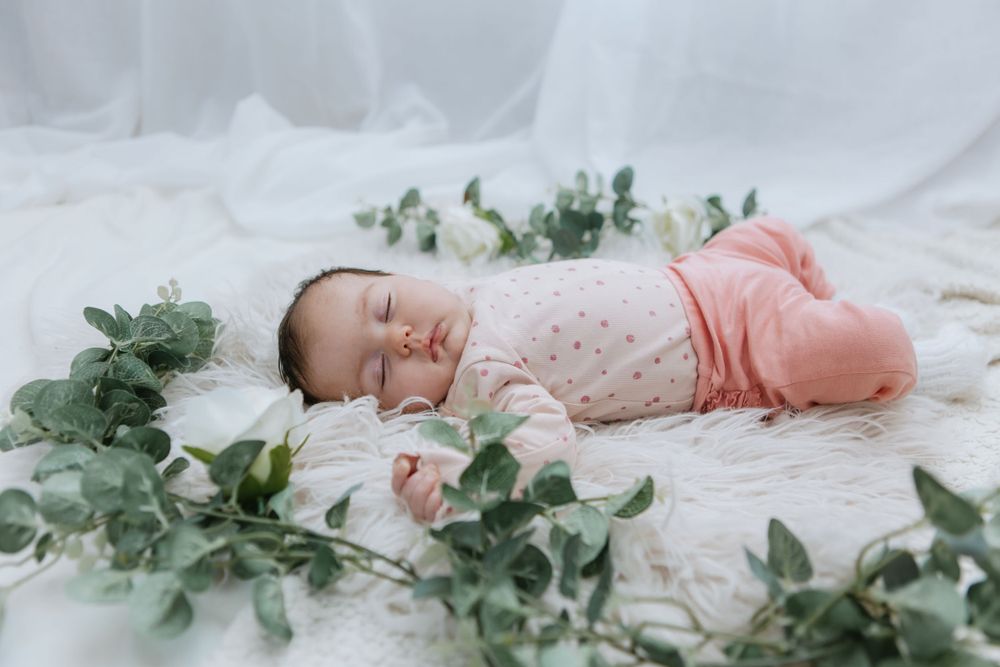 Peaceful sleeping newborn wearing pink outfit lies on white blanket surrounded by delicate green foliage.