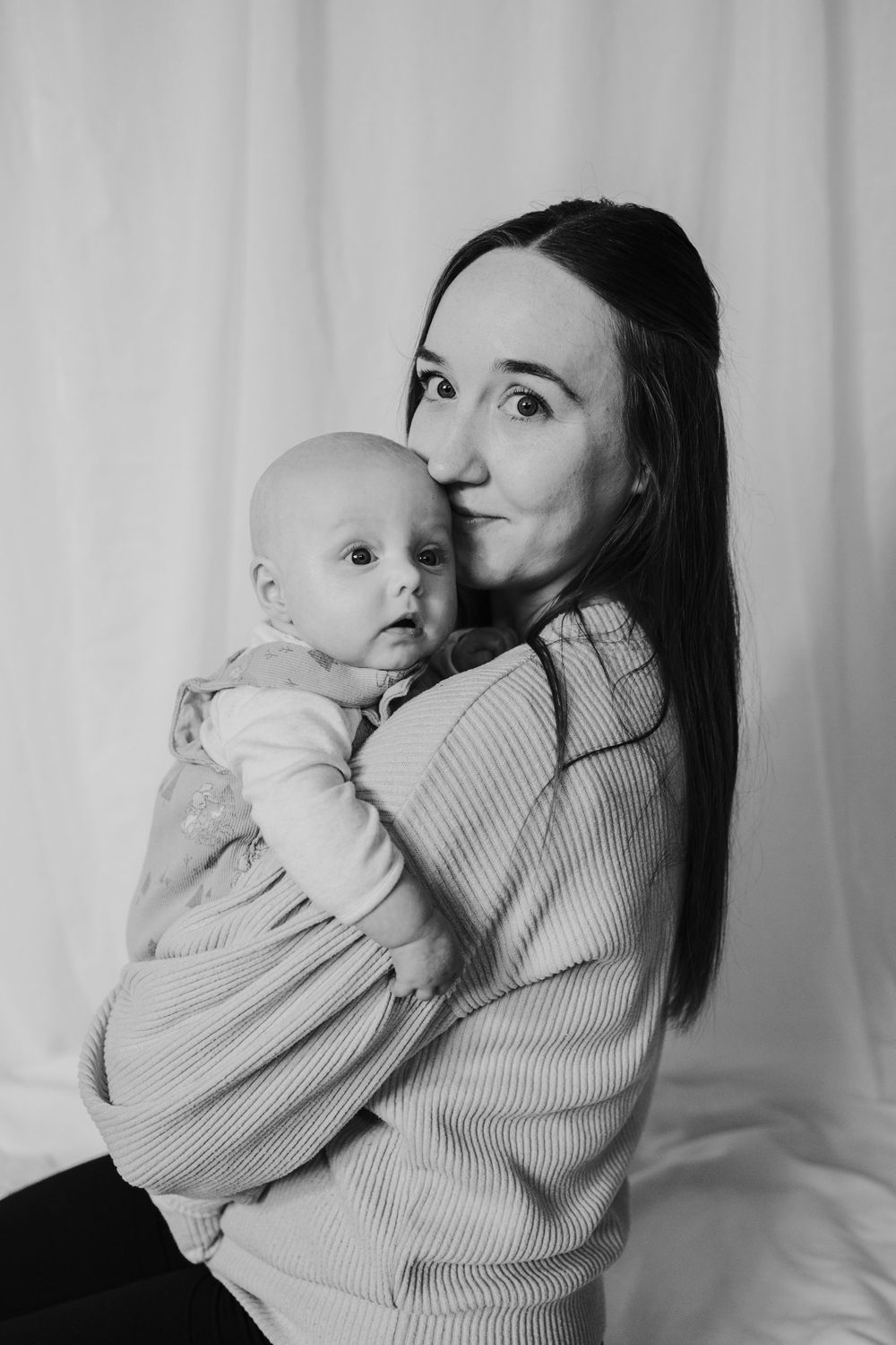 A black and white portrait shows a mother in a knit sweater embracing her baby in matching outfits against a light backdrop.