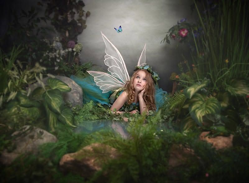 A young girl dressed as a fairy with wings and floral crown lies among lush green plants in an enchanted forest setting.