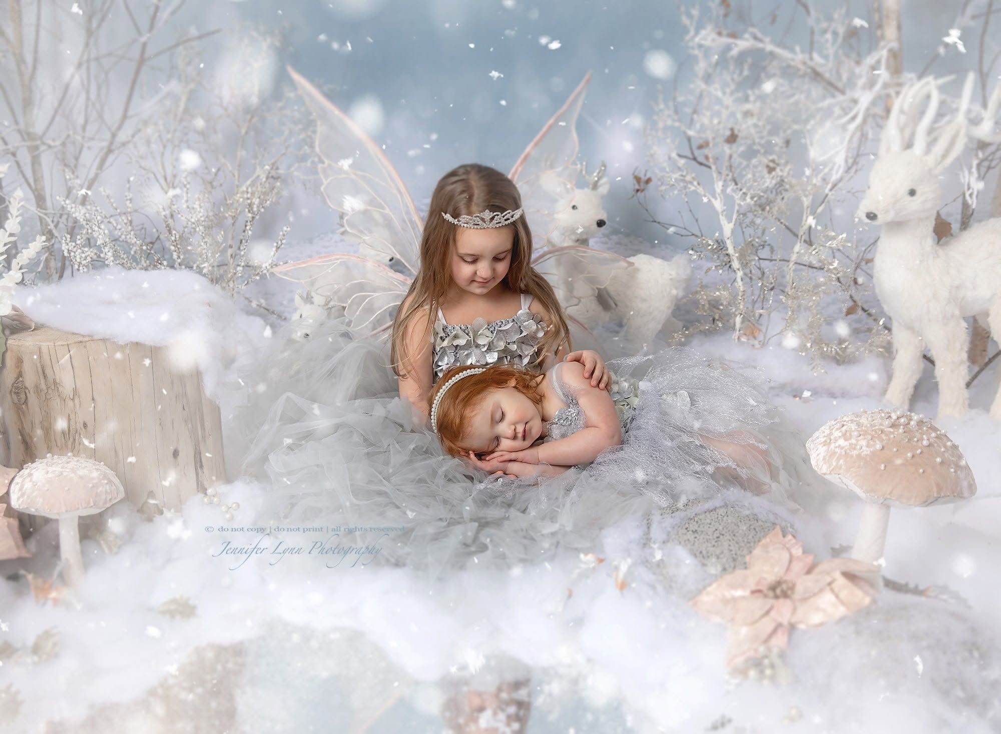 A young girl dressed as a fairy in a silver gown holds a sleeping baby in a magical winter wonderland scene.