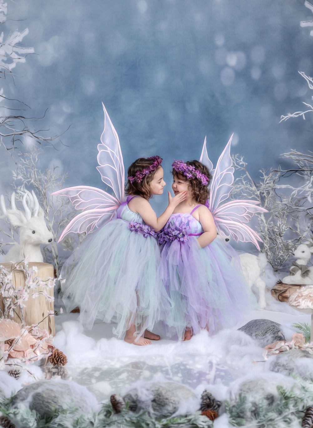 Two little girls in fairy costumes with wings and floral crowns pose together in a magical winter wonderland setting.