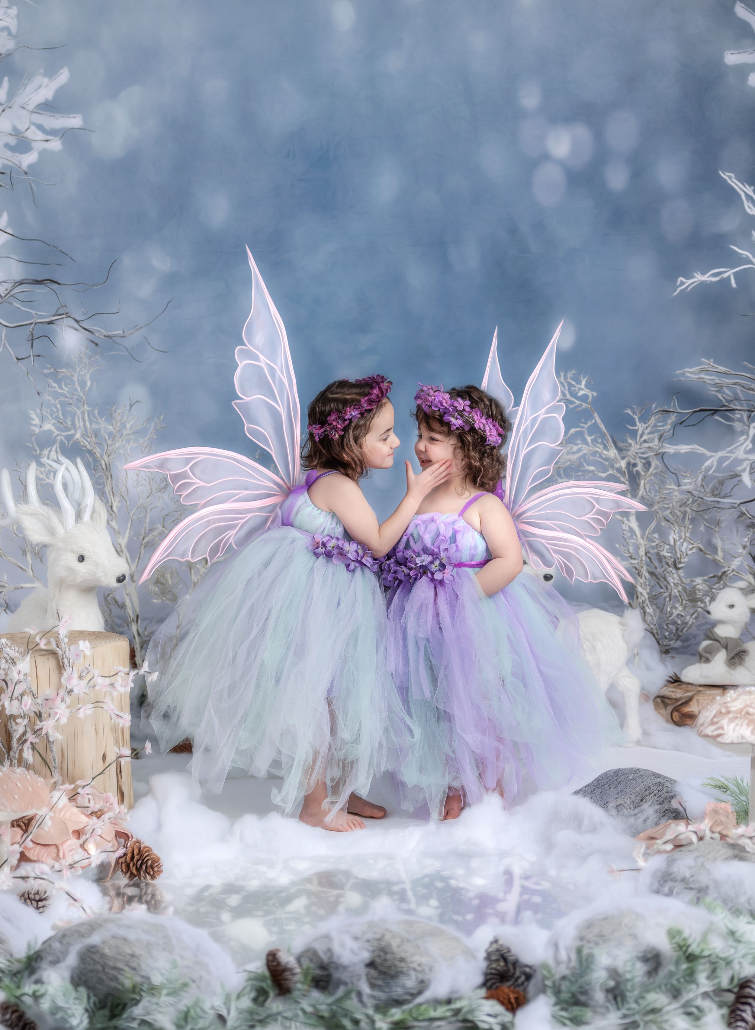 Two little girls in fairy costumes with wings and floral crowns pose together in a magical winter wonderland setting.