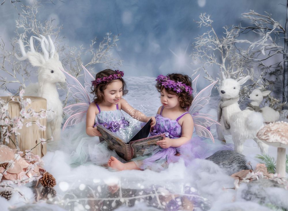 Two little girls dressed as fairies with butterfly wings and floral crowns read a book in a magical winter wonderland scene.
