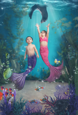 Two children dressed as mermaids underwater with colorful fish, coral, and a dolphin in a fantasy ocean scene.