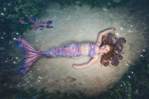 A young girl dressed as a mermaid with a colorful tail lies on a sandy ocean floor surrounded by fish and bubbles.