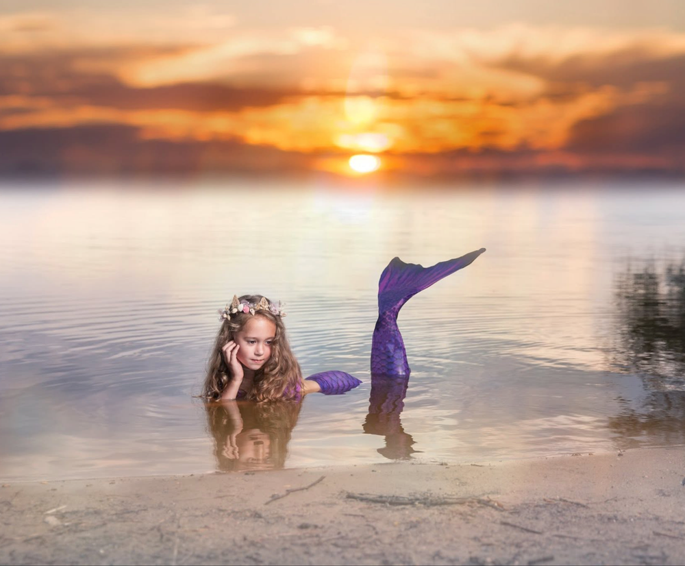 A young girl dressed as a mermaid with a purple tail poses in shallow water at sunset.
