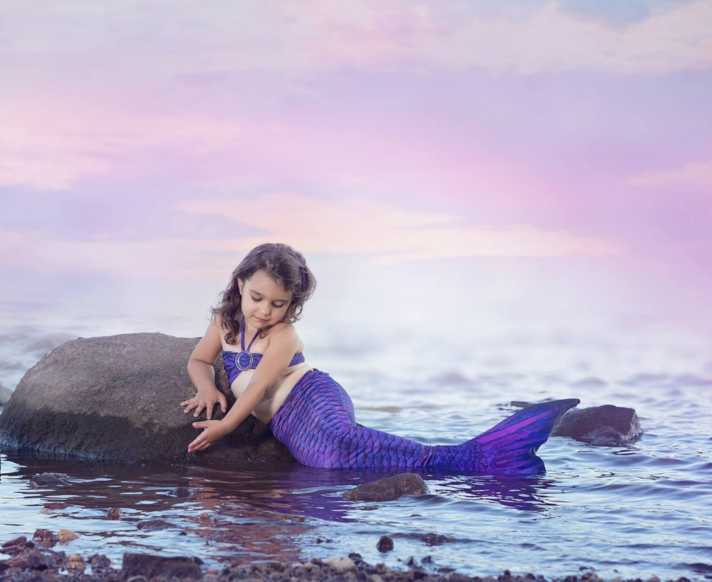 A young girl dressed as a mermaid with a purple tail rests on a rock in the ocean at sunset.