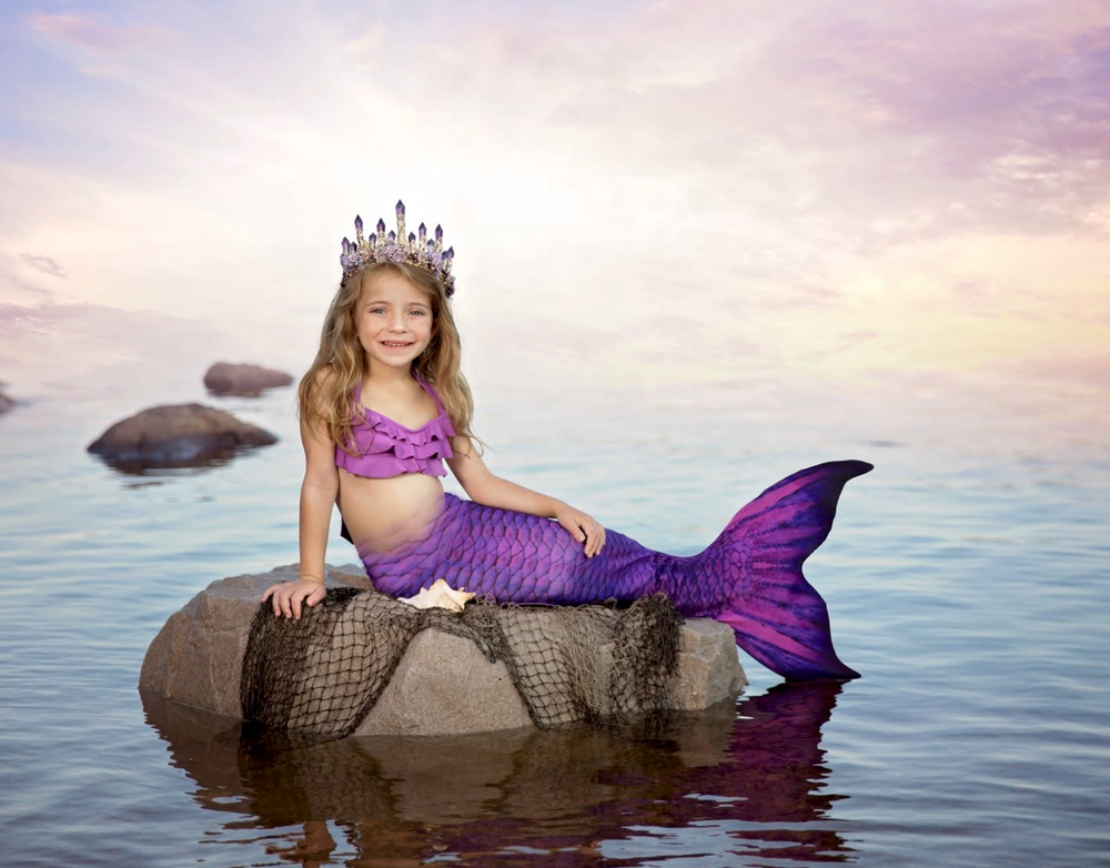 Young girl dressed as a mermaid with purple tail and crown sits on rocks in calm ocean water at sunset.