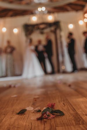 A fallen flower on a wooden floor with a blurred wedding ceremony and string lights in background.