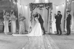Black and white wedding photo of couple sharing first kiss at rustic barn venue with floral arch and wedding party.