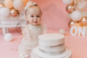 Smiling baby girl in white lace dress at first birthday cake smash with pink and gold balloon backdrop.