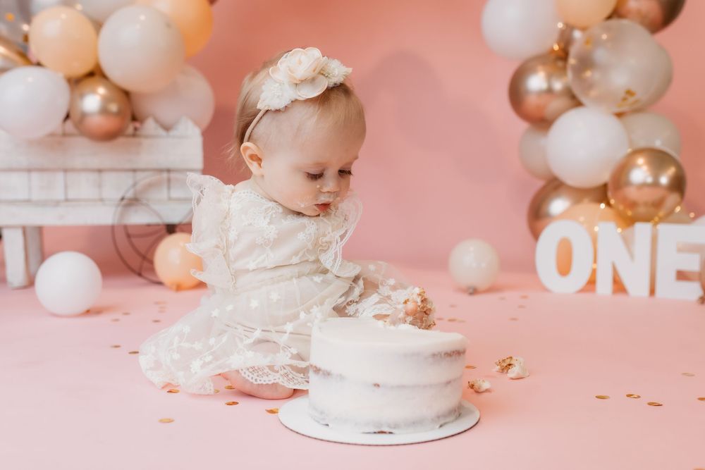 Baby girl in white dress smashes first birthday cake surrounded by gold and pink balloons spelling ONE.