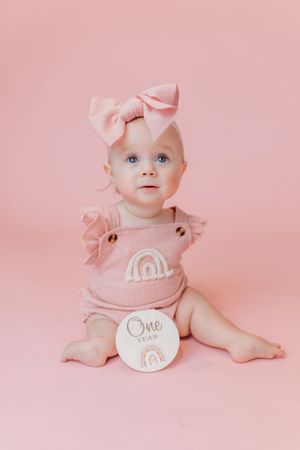 Adorable baby girl in pink romper and bow headband sits with a One Year milestone disc on pink background.