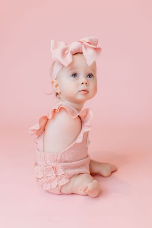 Adorable baby girl in pink ruffle romper and bow headband sitting on pink background.