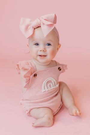 Adorable baby girl in pink romper with rainbow embroidery and bow headband sitting on pink background.