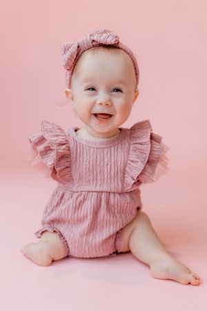Smiling baby girl in pink ruffle romper and bow headband sitting against a pink background.