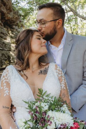 A groom in a gray suit kisses his bride on the cheek as she smiles holding a white and purple bouquet outdoors.