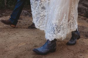 Bride in lace wedding gown wearing black cowboy boots standing beside groom in blue jeans and brown western boots.
