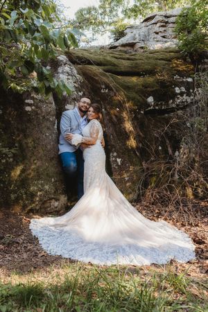Bride and groom embracing against a mossy rock formation outdoors, bride wearing a lace gown with long train.