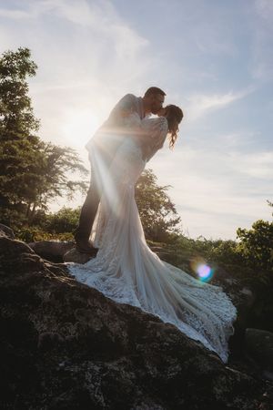 Bride and groom share a romantic kiss on rocky terrain at sunset, wedding dress trailing elegantly.