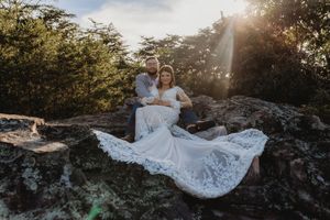 Couple in white lace maternity gown sitting on rocks at sunset surrounded by lush green trees.