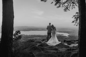 Black and white photo of bride and groom standing on rocky overlook with panoramic lake and valley views.