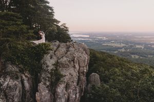 Bride and groom sit atop a stunning rocky cliff overlooking a lush green valley at sunset during their outdoor wedding.
