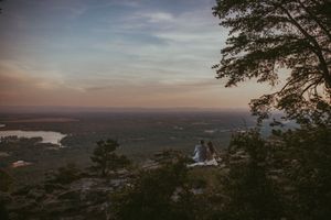 Two people sit on a rocky overlook enjoying a stunning sunset panoramic view of forests and lakes below.