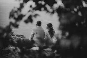 Black and white photo of a bride and groom sitting together on a cliff, gazing at the ocean view.