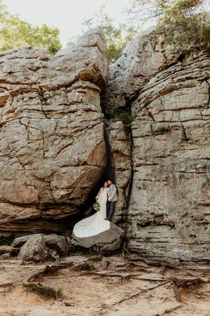 Bride and groom standing in a rocky canyon crevice, bride in flowing white gown, outdoor wedding photo.